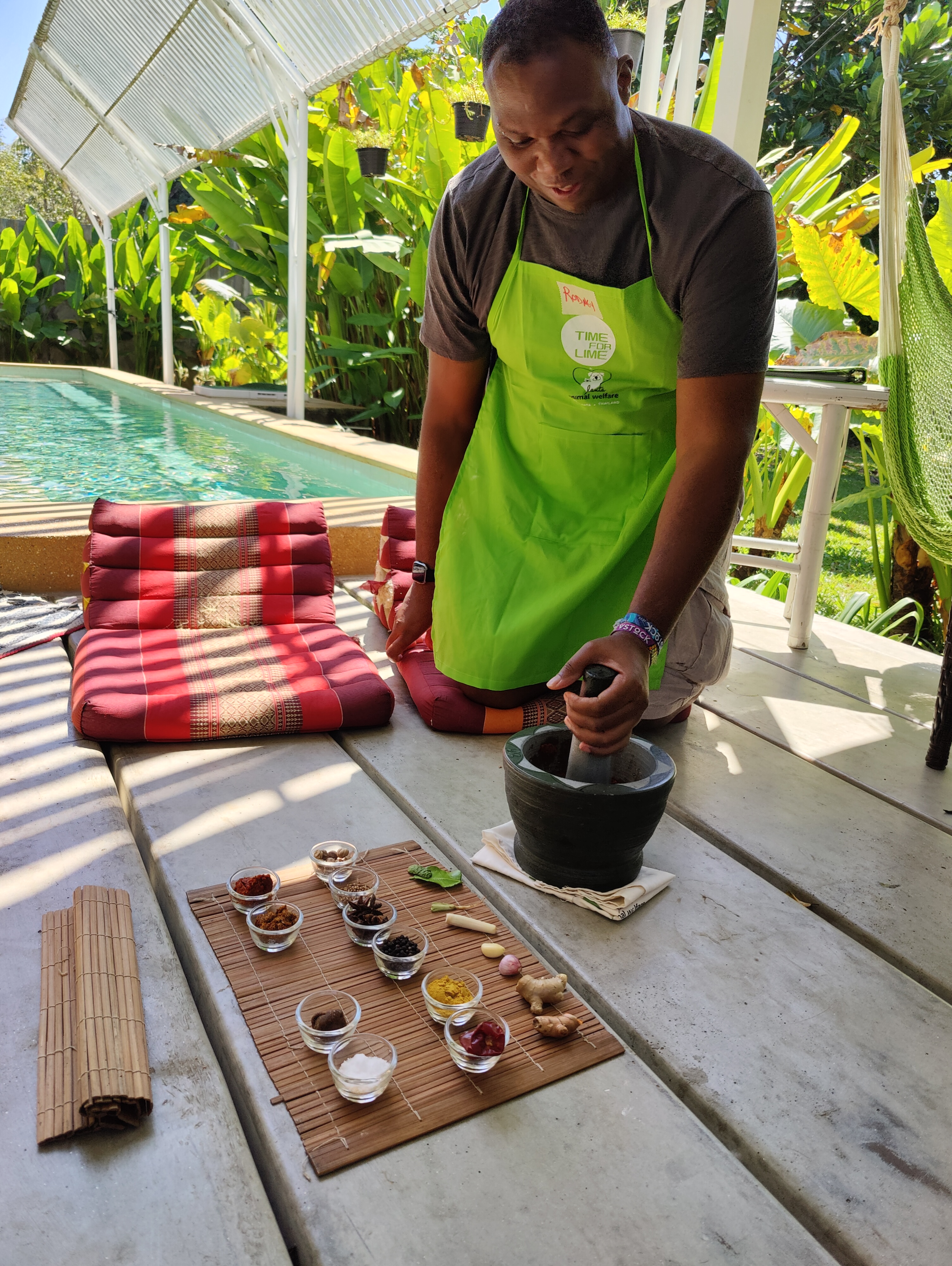 Rodney in green Time for Lime apron, working a mortar and pestle, poolside garden